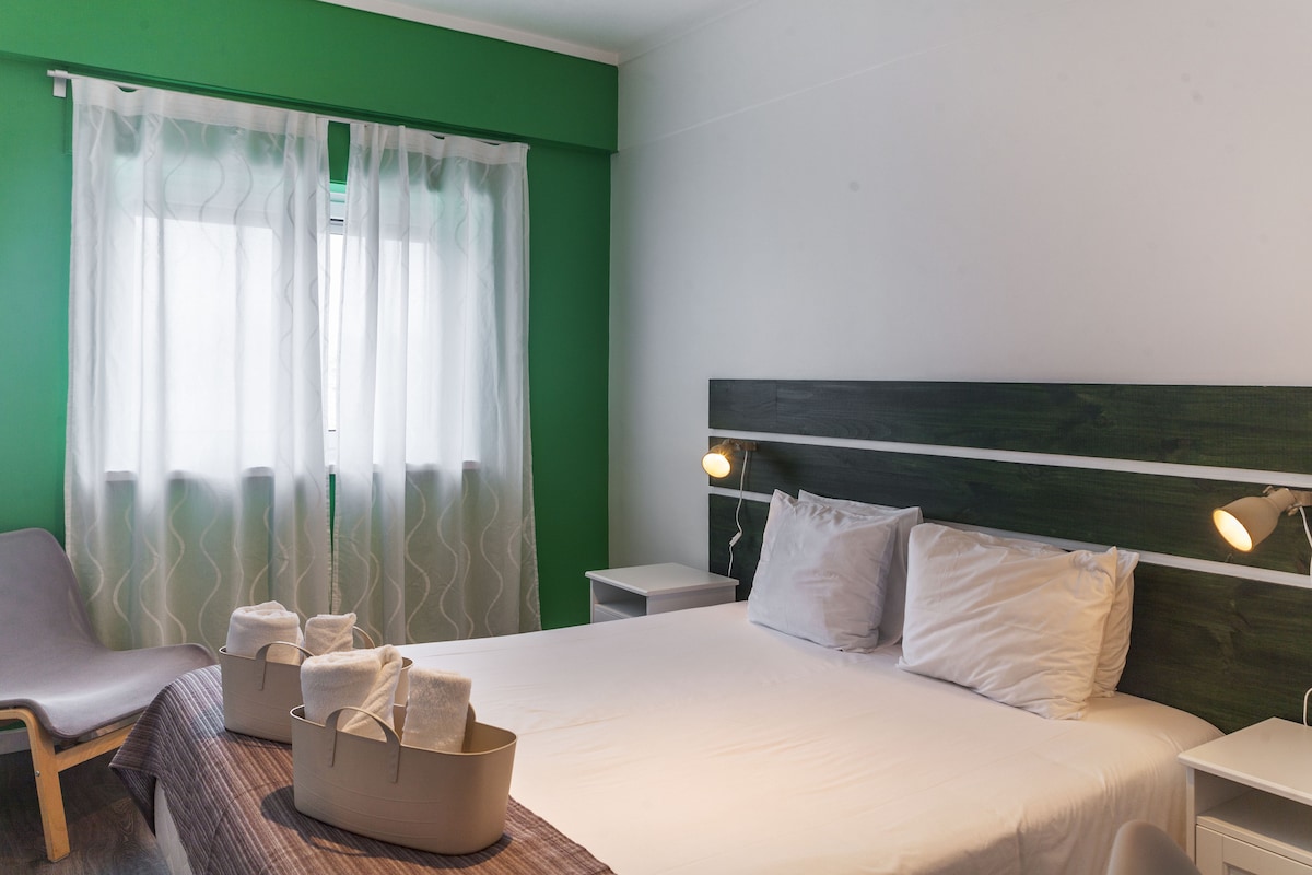 A spacious room is presented with a double bed featuring white linens and a textured throw. Soft lighting is provided by wall-mounted lamps. A minimalist chair and bedside tables complement the green accent wall, while sheer curtains allow natural light to filter in.