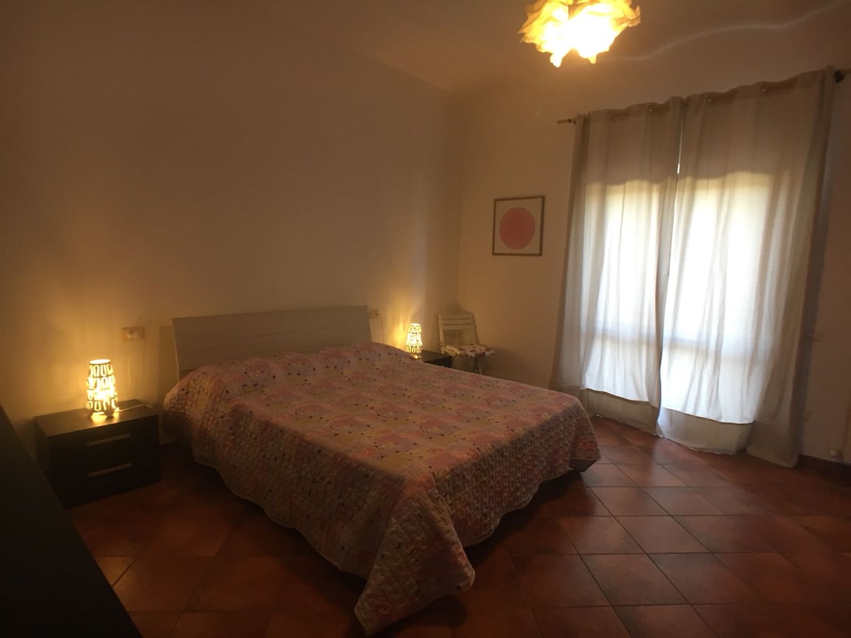 A spacious bedroom is presented, featuring a large bed covered with a patterned quilt. Soft lighting is provided by bedside lamps on either side, while natural light filters through sheer curtains, creating a calm environment. The terracotta tiles add warmth to the space.