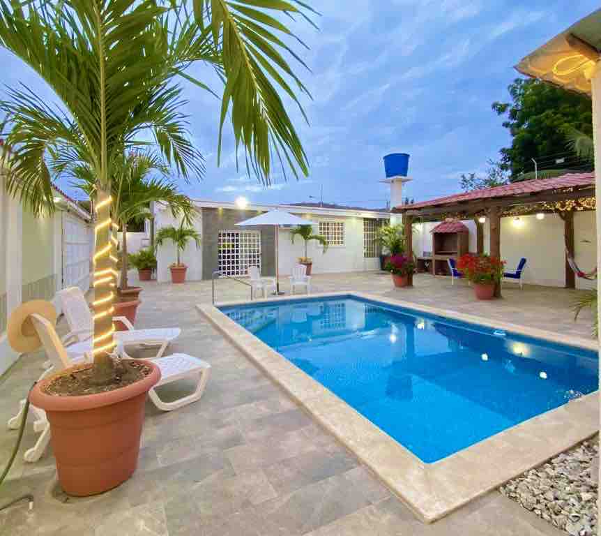 A private outdoor area features a clear blue swimming pool surrounded by patio tiles. Sun loungers are positioned nearby, complemented by potted plants and shades. Soft evening light casts a calm ambiance over the space, enhancing its inviting atmosphere.