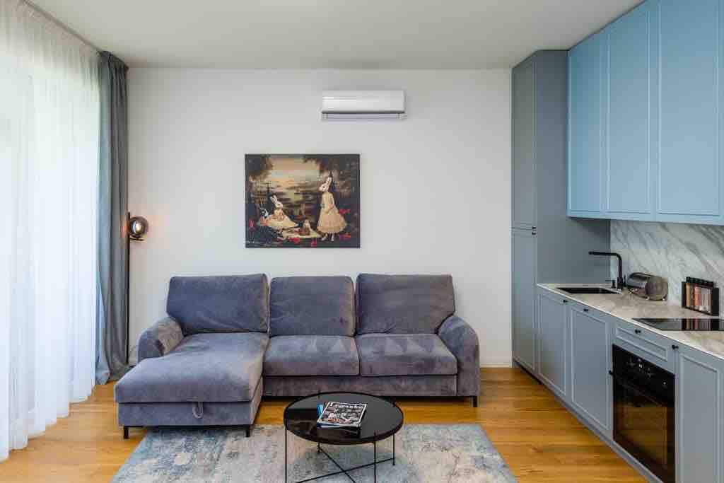 A living area is presented with a large gray sectional sofa positioned against a wall. A black coffee table rests in front, and a painting depicting a serene scene is hung above the seating. Natural light filters through sheer curtains, illuminating the wooden floor.