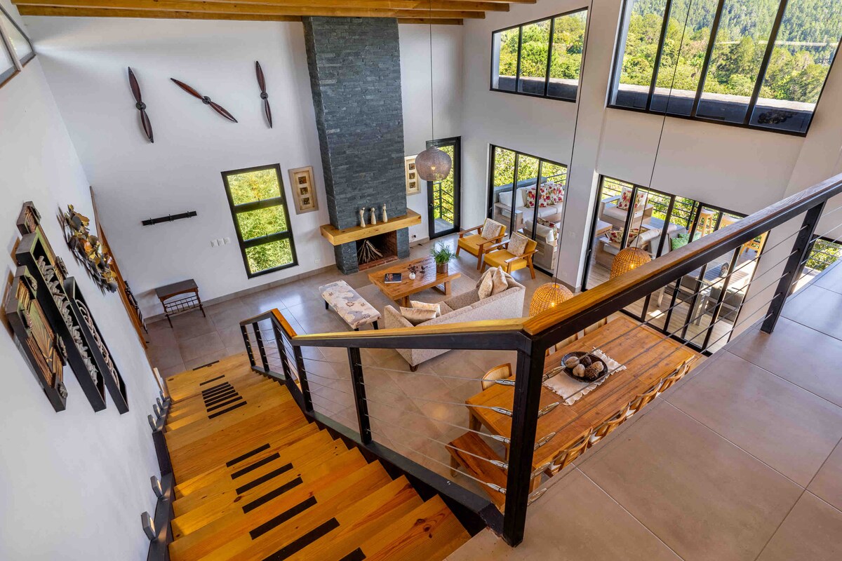 A spacious living area features high ceilings accented by wooden beams. Large windows allow natural light to illuminate the open layout, which includes a comfortable seating arrangement and a central fireplace. A staircase with wooden steps leads to the upper levels, showcasing the modern architecture.