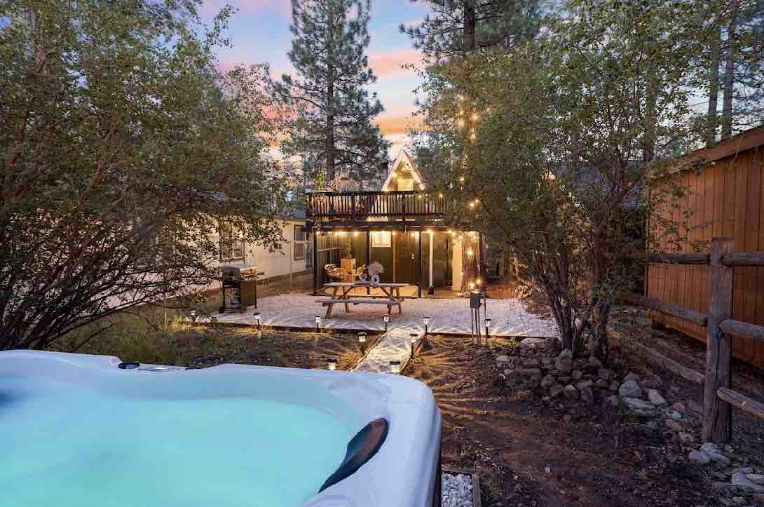 A spacious outdoor area is visible, featuring a new hot tub in the foreground. String lights create a warm ambiance around a wooden picnic table, surrounded by natural landscaping. The A-frame structure is partially seen in the background, framed by tall trees as dusk sets in.