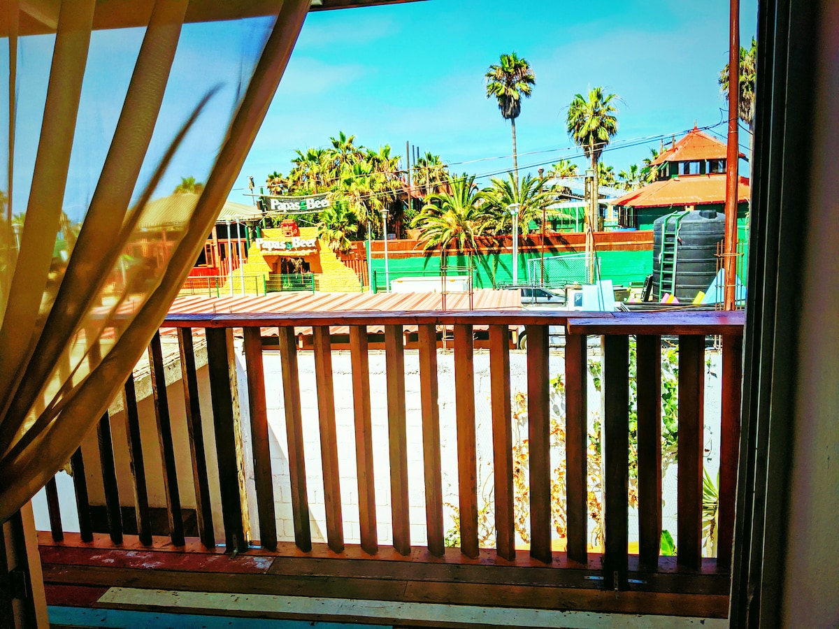 A view from a balcony showcases palm trees and vibrant greenery in the foreground, while the lively Papas & Beers club is visible nearby. The clear blue sky adds brightness to the scene, with sunlight streaming onto the wooden railing.
