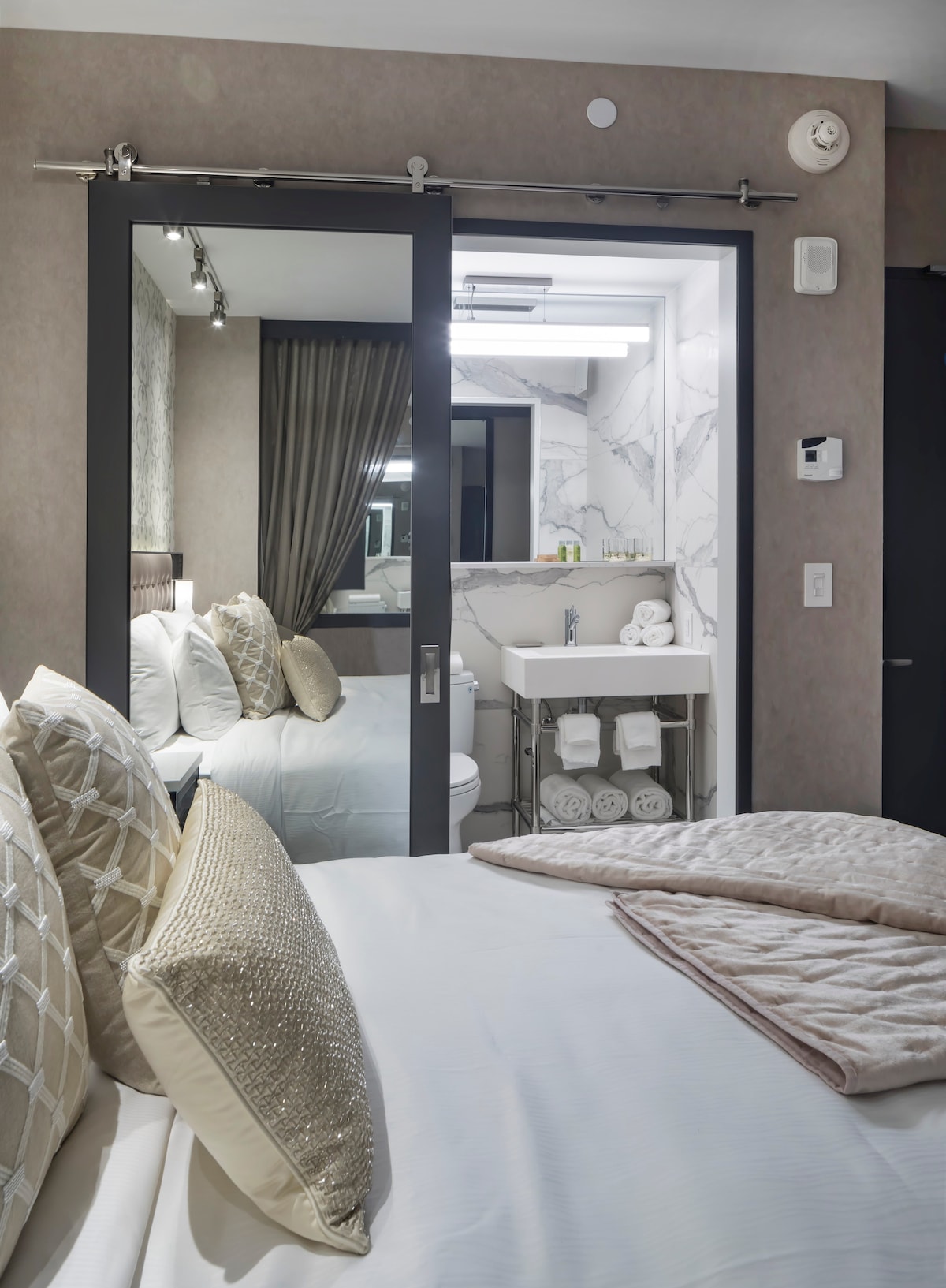 A stylish bedroom displays a spacious bed adorned with textured pillows and a soft throw. A glass door opens to a modern bathroom featuring marble accents, neatly arranged towels, and a well-lit vanity area.