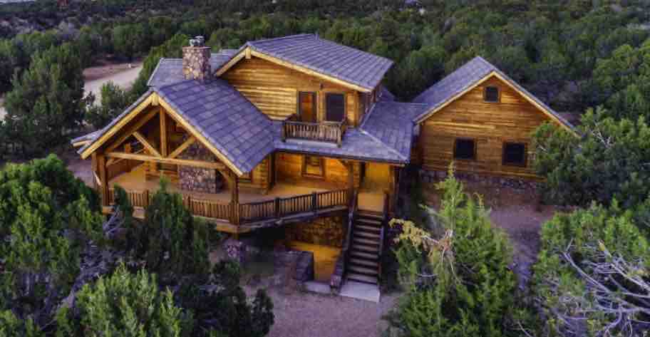 The spacious log cabin features a gabled roof and expansive decks with wooden railings. It is surrounded by lush juniper trees, emphasizing its secluded location. Soft lighting emanates from the windows, suggesting a welcoming ambiance in the evening.