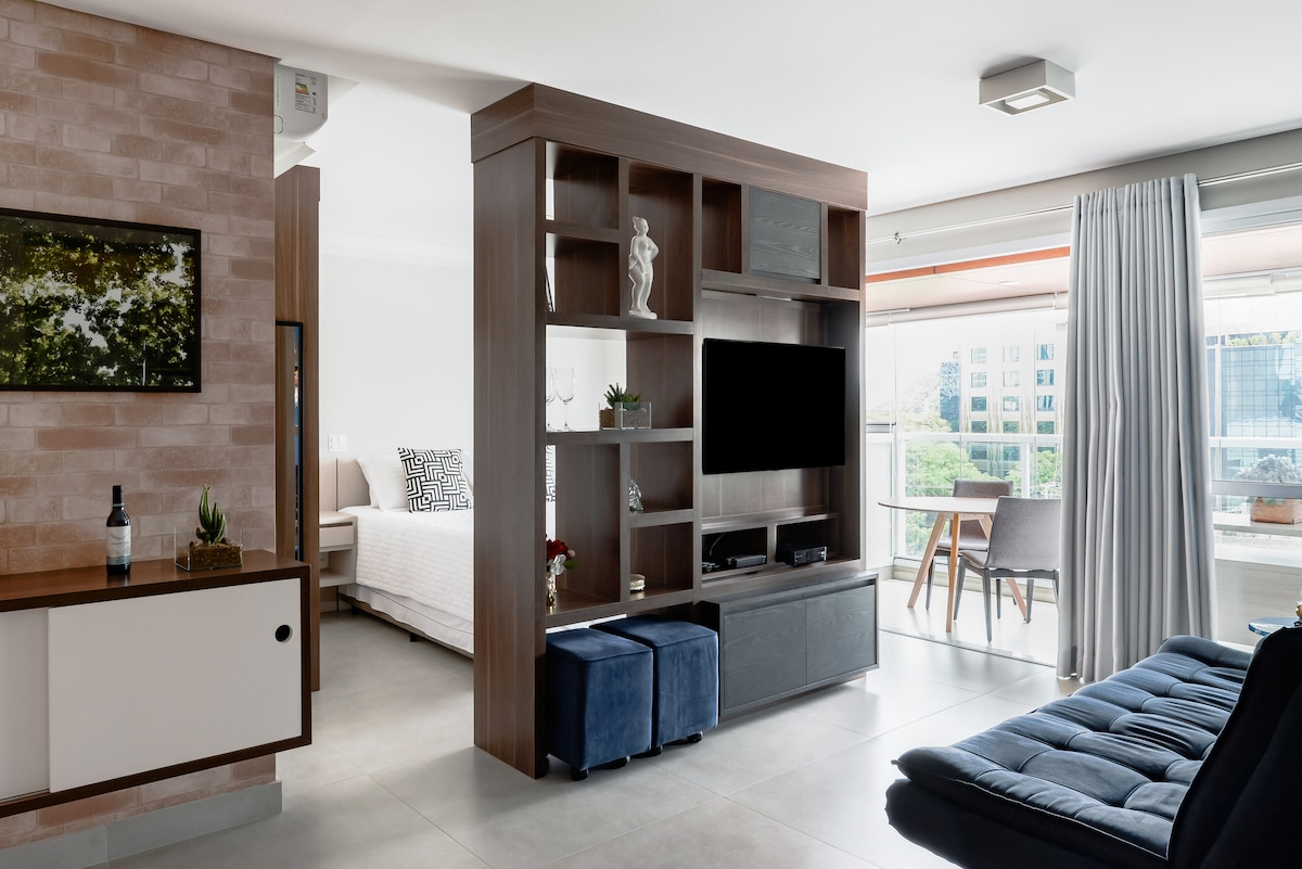 A modern studio space is visible, featuring a stylish shelving unit with decorative items and a mounted television. The area includes a cozy blue sofa, a dining table with chairs, and large windows allowing natural light. The décor is minimal and contemporary.