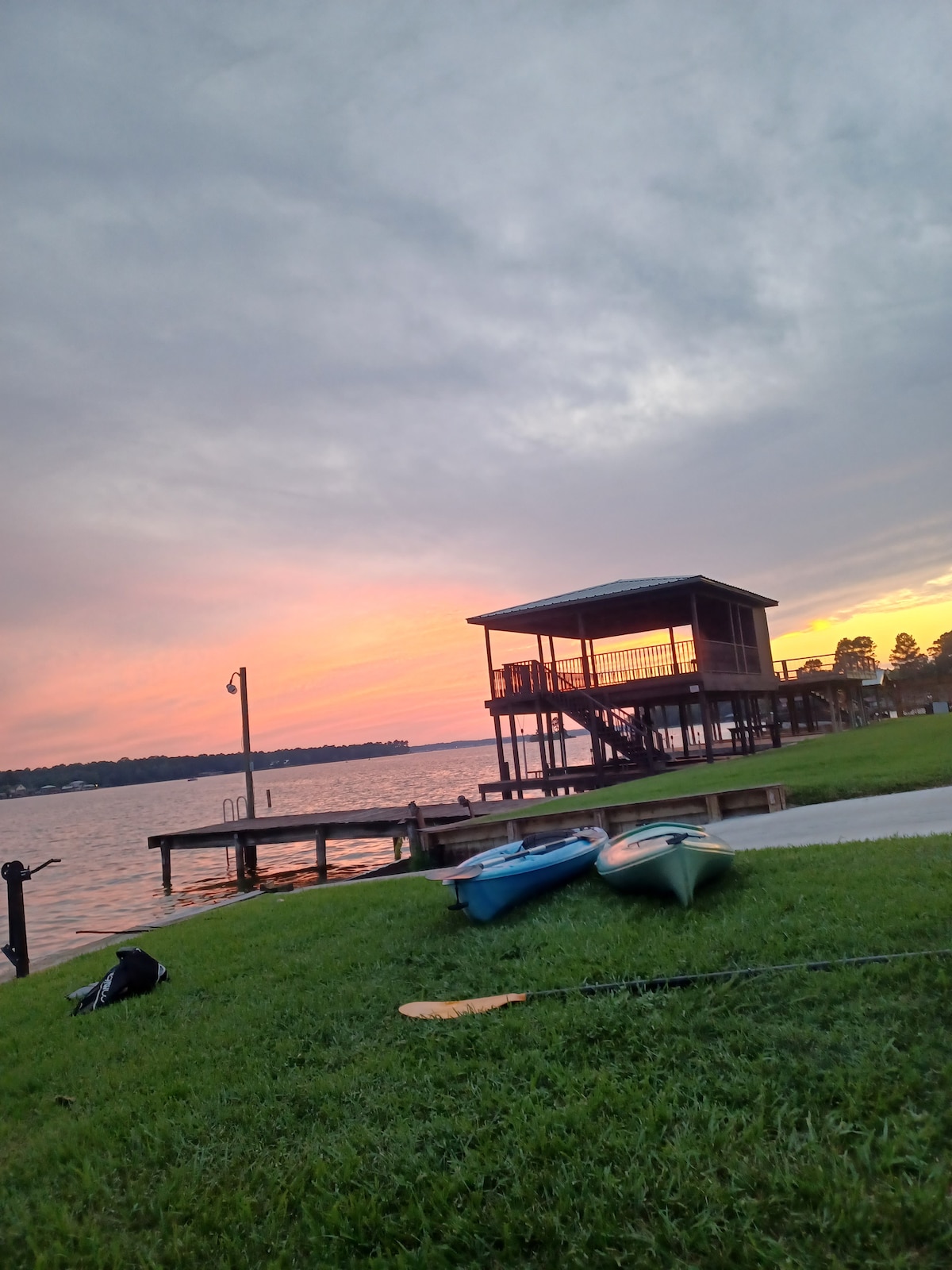A serene lakeside view showcases a wooden dock and a boat house under a sunset sky. Two kayaks rest on the lush green grass, while soft colors blend in the horizon. The calming water reflects the fading light, enhancing the peaceful atmosphere.