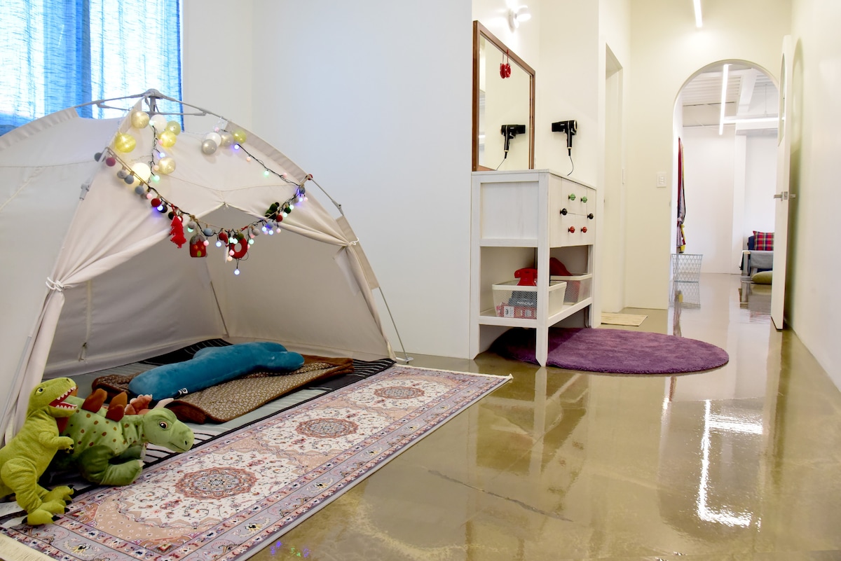 A cozy play area features a fabric tent adorned with decorative lights. Plush toys, including dinosaurs, are positioned on a patterned rug alongside a sleeping mat. A mirrored dresser with colorful drawer handles is visible in the background, complemented by a circular purple rug on the polished floor.