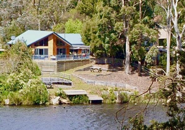 A wooden house with a blue roof overlooks a serene waterfront. A deck extends from the house, offering outdoor seating options. Lush greenery and trees surround the property, with pathways leading to a picnic area by the water's edge.