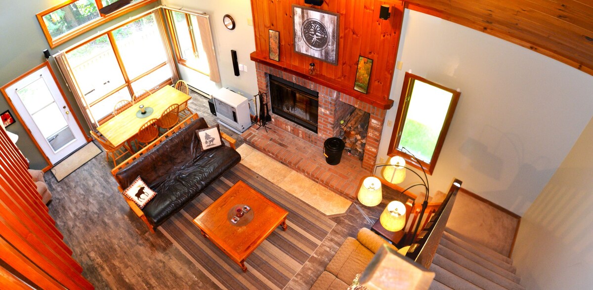 The view captures a spacious living area featuring a stone fireplace and comfortable seating arrangements. Natural light floods the room through large windows. A dining table is positioned adjacent to the kitchen, creating an open and inviting space. Stairs lead to the upper level.