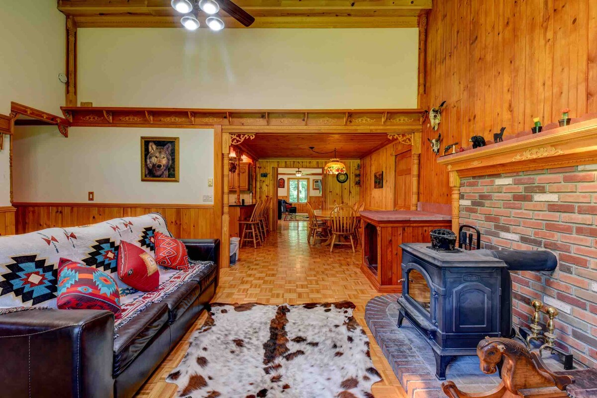 A welcoming living area is furnished with a dark leather sofa adorned with decorative pillows. A wood-burning stove is positioned beside a rustic stone fireplace. The space features wooden walls and a patterned rug, leading toward an open dining area visible in the background.