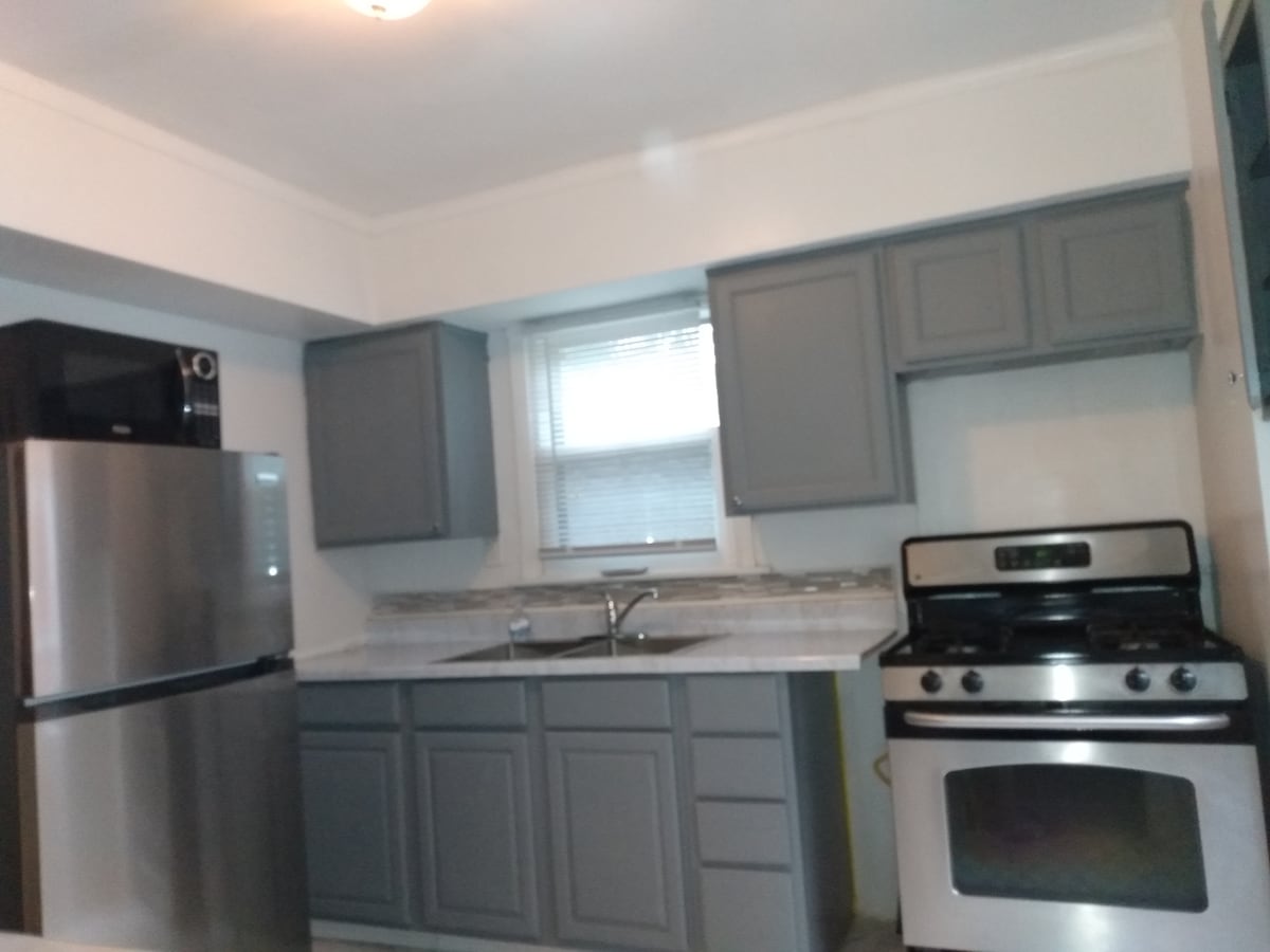 The kitchen is equipped with modern stainless steel appliances, including a refrigerator and a gas stove. Gray cabinets and a marble countertop create a clean and functional space. Natural light enters through the window above the sink.