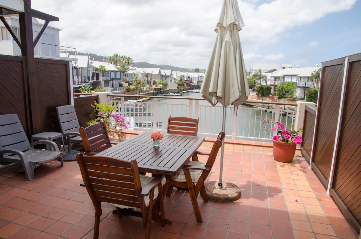 A spacious balcony is featured, furnished with a wooden dining table surrounded by chairs. An umbrella provides shade, and colorful potted flowers enhance the space. The view of the water and nearby buildings complements the tranquil outdoor setting.
