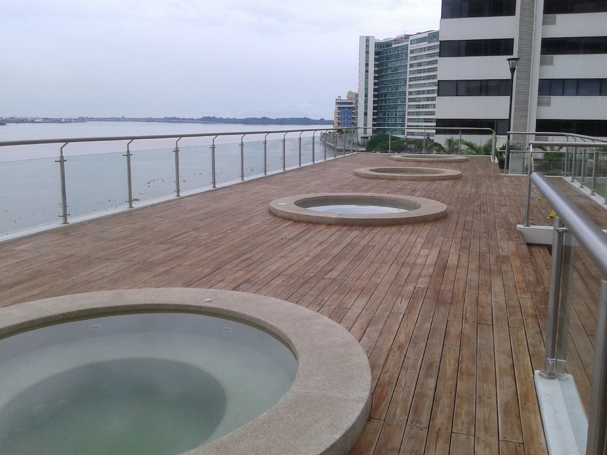 A spacious outdoor area features multiple circular jacuzzis situated on a wooden deck, offering views of the river. The surrounding railing ensures safety while allowing unhindered access to the scenic waterfront. Buildings are visible in the background, enhancing the urban setting.