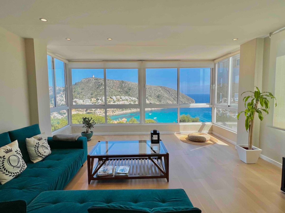 A modern living area features floor-to-ceiling windows showcasing a stunning view of the bay and surrounding mountains. A plush green sofa is arranged around a wooden coffee table, while a potted plant adds a touch of nature to the space. Natural light enhances the bright interior.