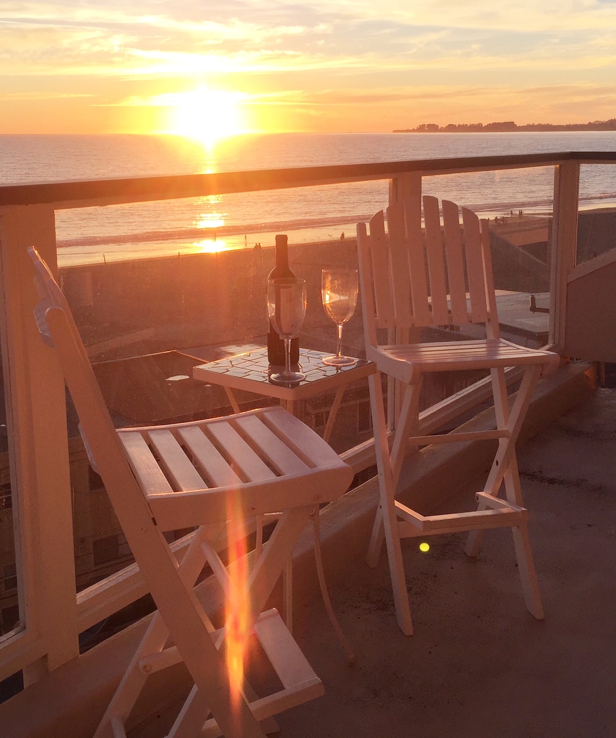 A small balcony is furnished with two white folding chairs and a matching table. A bottle of wine and a glass sit on the table, illuminated by the warm glow of the setting sun over the Pacific Ocean.