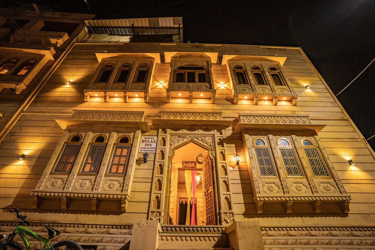 An intricately designed building exterior is lit softly by warm lights. Decorative carvings adorn the walls, highlighting the architectural details. Multiple windows feature ornate lattice work, contributing to the distinctive character of the structure. A welcoming entrance is visible at the center.