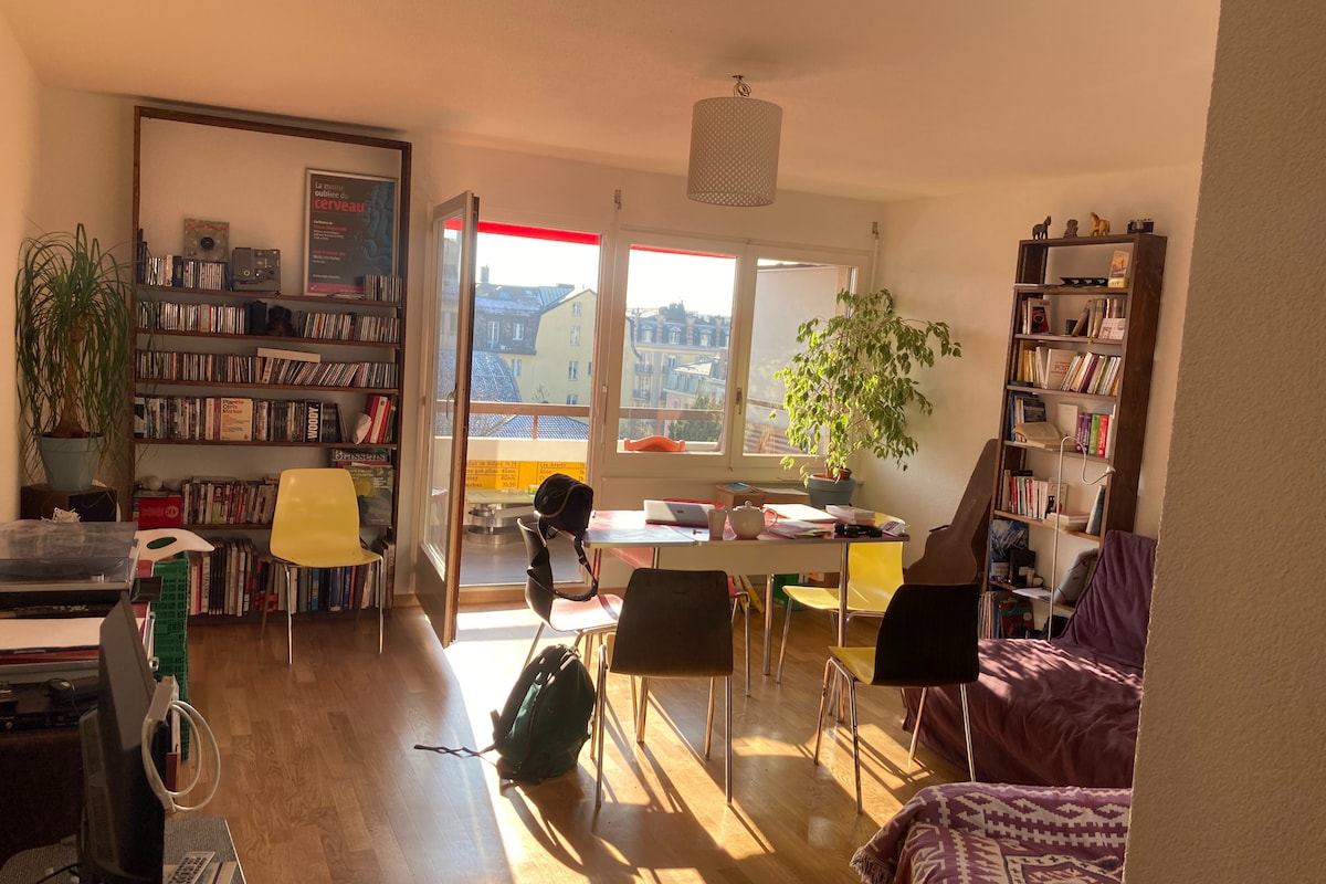 A bright living area features large windows allowing natural light to fill the space. A dining table is surrounded by several chairs, while a cozy seating area includes a relaxed chair and a bookshelf filled with various items. A leafy plant adds a touch of greenery.