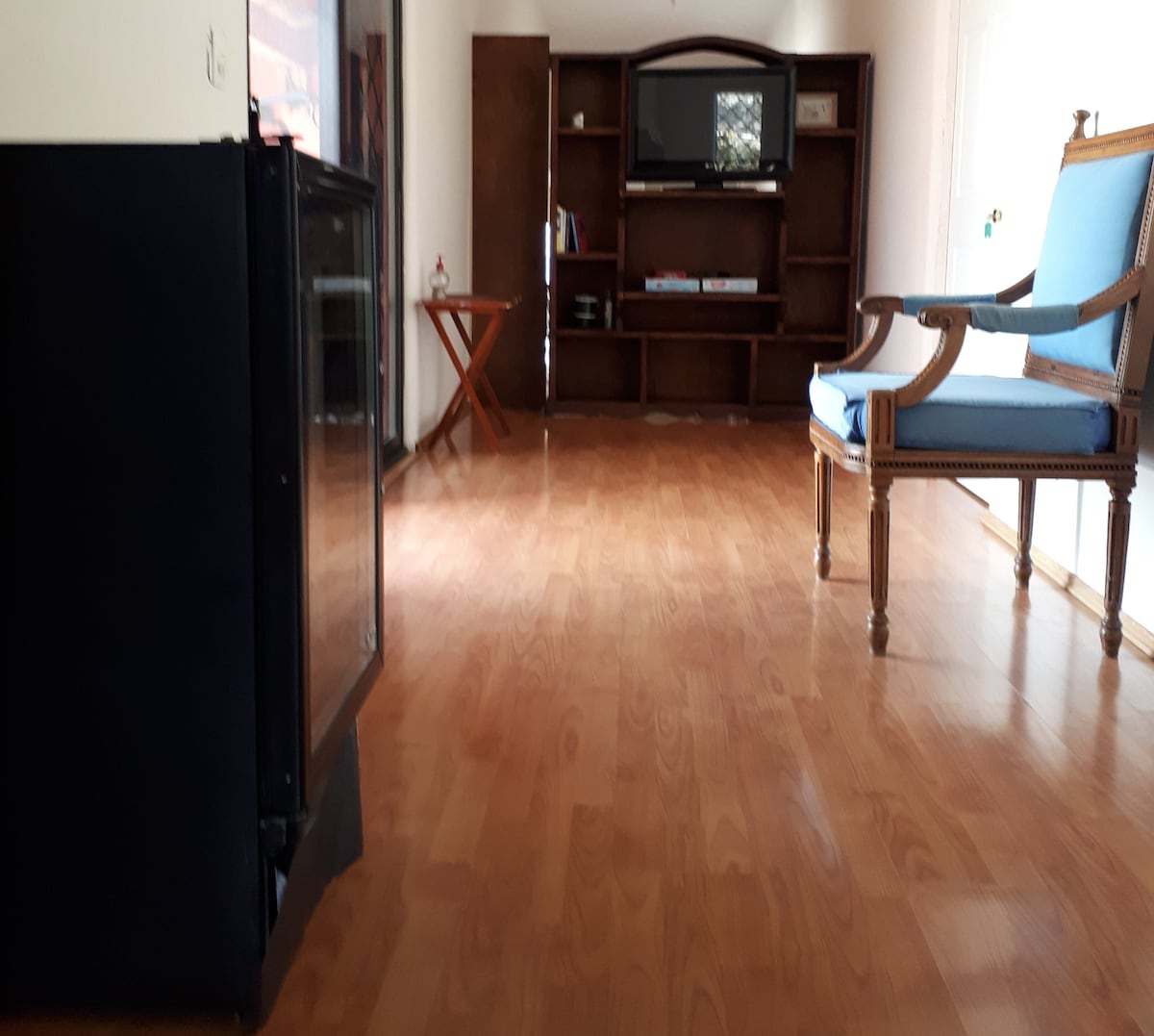 A spacious living area features polished wooden flooring. A stylish blue upholstered chair and a wooden side table are positioned nearby. A wooden shelf holds a television and various items, with ample natural light illuminating the room.