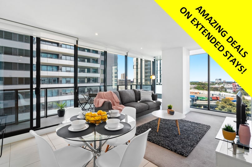 A bright living area is featured, showcasing large floor-to-ceiling windows that show scenic views of the surrounding city. A comfortable grey sofa is positioned beside a glass coffee table adorned with a bowl of fruit. A dining table, set for four, is located nearby on a textured rug.