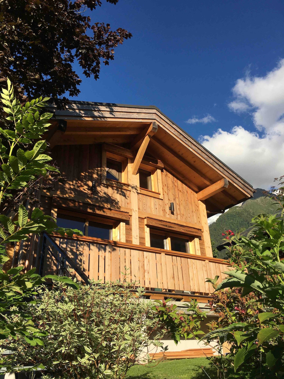 The charming wooden chalet is set against a clear blue sky, showcasing its rustic architecture with a sloped roof and numerous windows. A small balcony is visible, framed by greenery and flowering plants, emphasizing the connection with the natural surroundings.