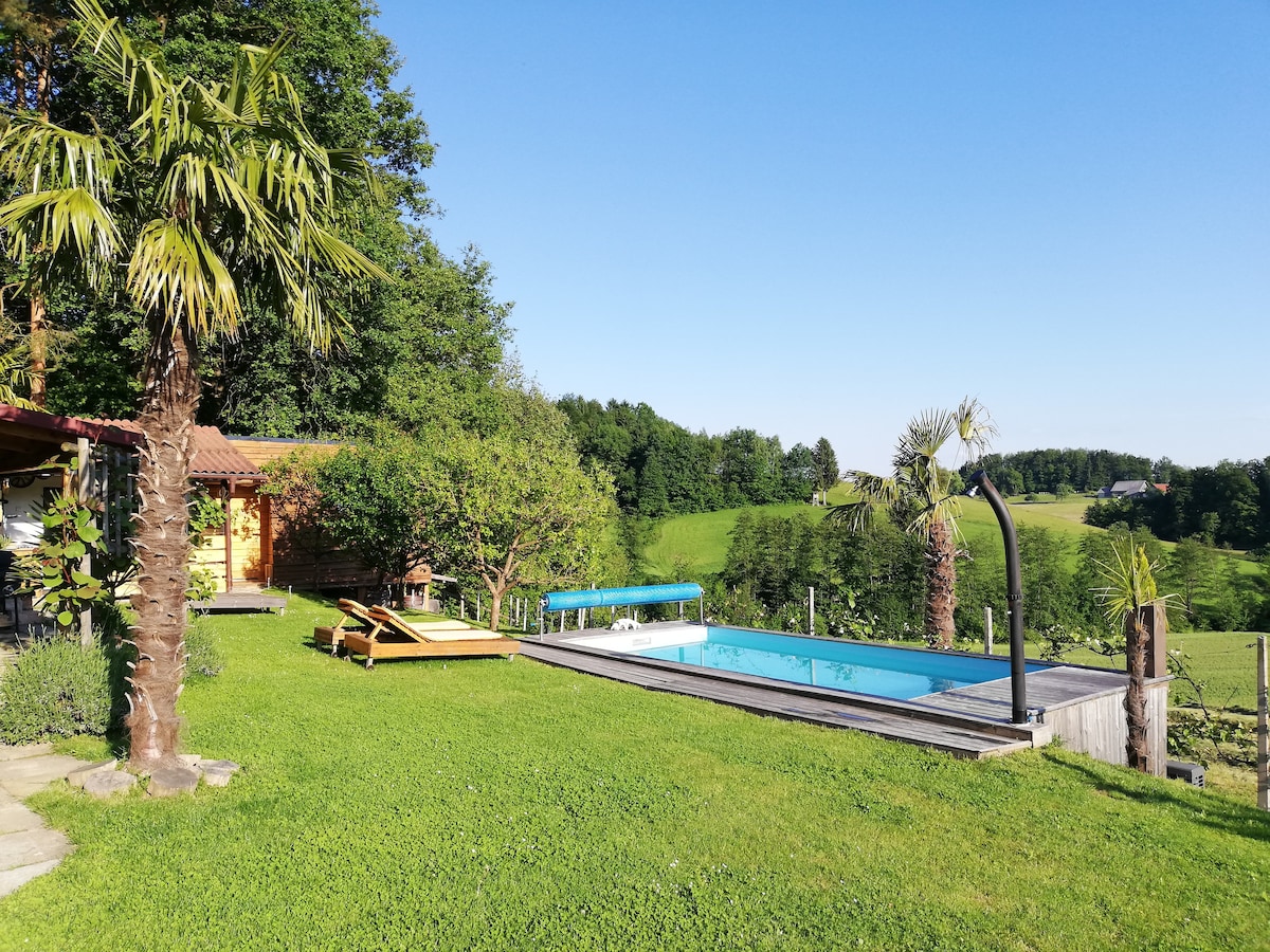 A sunlit outdoor area features a heated pool surrounded by well-maintained grass and palm trees. Sun loungers are positioned nearby, offering a space for relaxation. The backdrop consists of lush greenery and distant hills, contributing to a serene atmosphere.