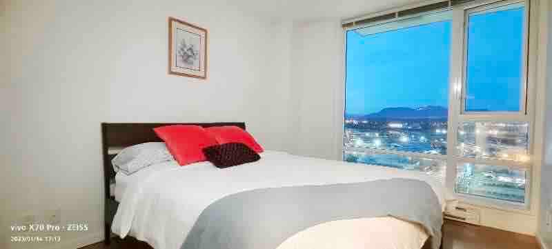A well-lit bedroom features a spacious bed adorned with a light grey blanket and decorative pillows. Large windows offer a view of the evening sky and city lights, enhancing the room's inviting atmosphere. A piece of wall art is hung above the bed.