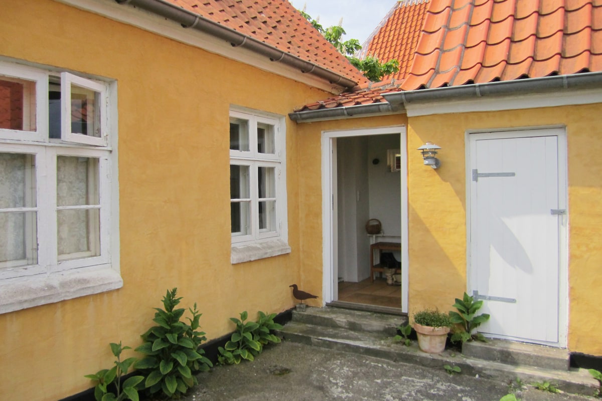 The charming exterior of a yellow house is framed by red tiled roofs. An inviting entrance, marked by a white door, offers access to the interior. Lush greenery and a small plant pot enhance the welcoming atmosphere of the pathway leading to the entrance.