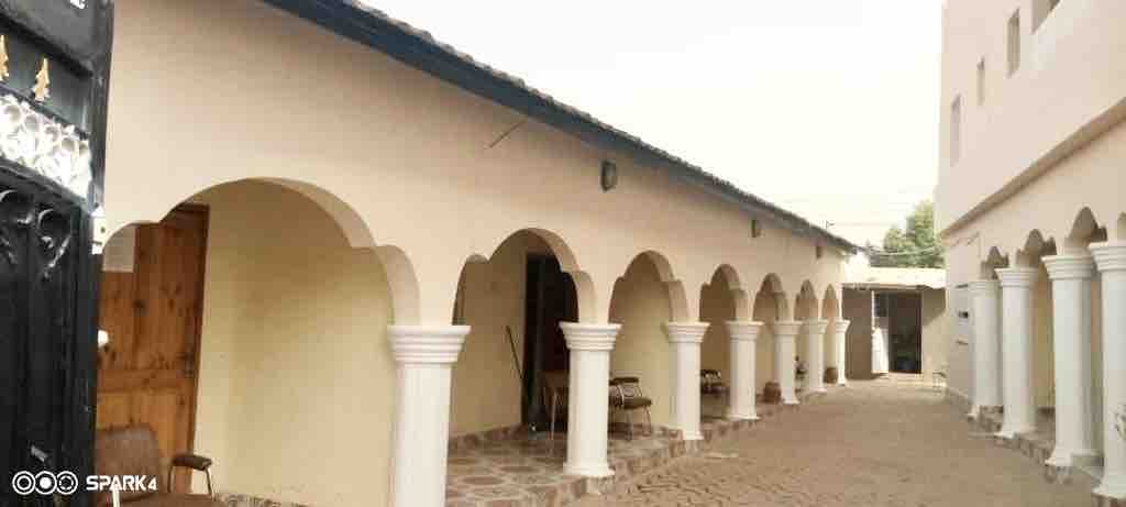 A walkway featuring arched columns and stone flooring is lined with wooden chairs. The structure's clean design and traditional elements reflect the local architecture. Natural light illuminates the corridor, contributing to a welcoming atmosphere.