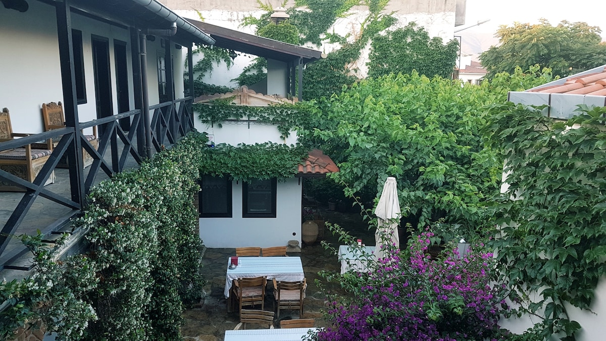 A tranquil courtyard is surrounded by greenery, featuring wooden tables and chairs beneath a canopy of vines. Flowering plants add vibrant color to the space, while a shaded umbrella provides comfort. The rustic architecture includes a mix of stone and wood elements.