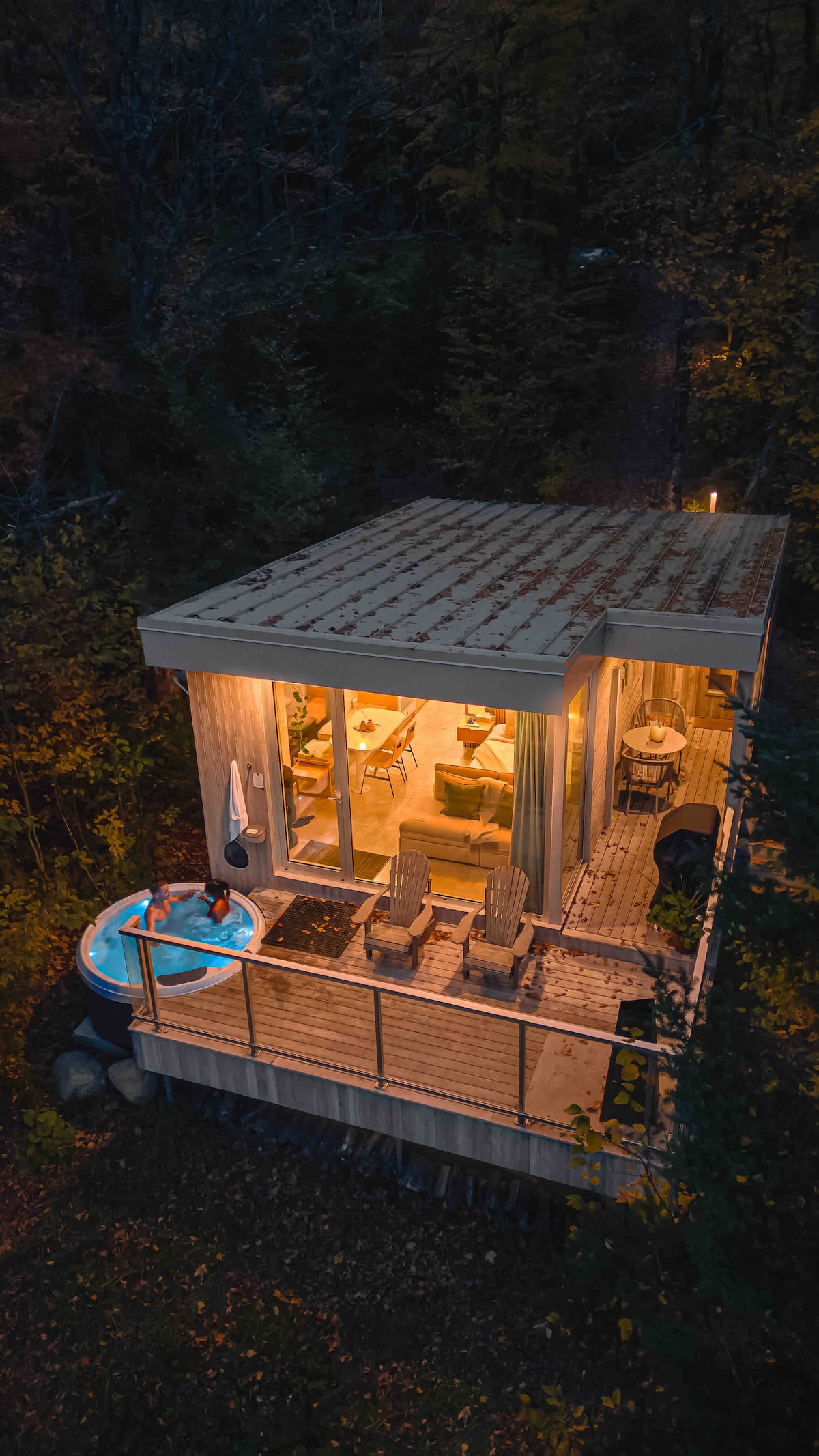 An aerial view showcases the chalet's modern exterior, highlighting its wooden deck and inviting hot tub. Warm light spills from the interior, illuminating a cozy seating arrangement inside. Surrounding trees create a natural, serene atmosphere, enhancing the feeling of privacy.