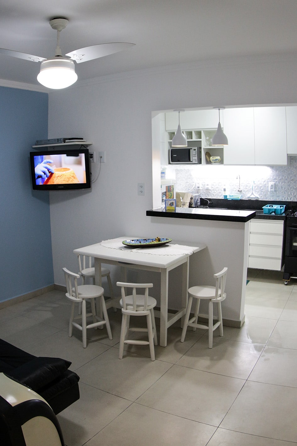The dining area features a small white table surrounded by four light-colored chairs. A wall-mounted television displays a program, while a ceiling fan provides airflow. A glimpse of the kitchen is visible, showcasing modern appliances and bright cabinetry.