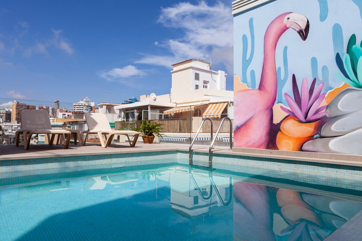 A rooftop pool is surrounded by comfortable loungers and a vibrant mural featuring a flamingo and succulent plants. The clear water reflects the blue sky, creating a serene environment for relaxation.