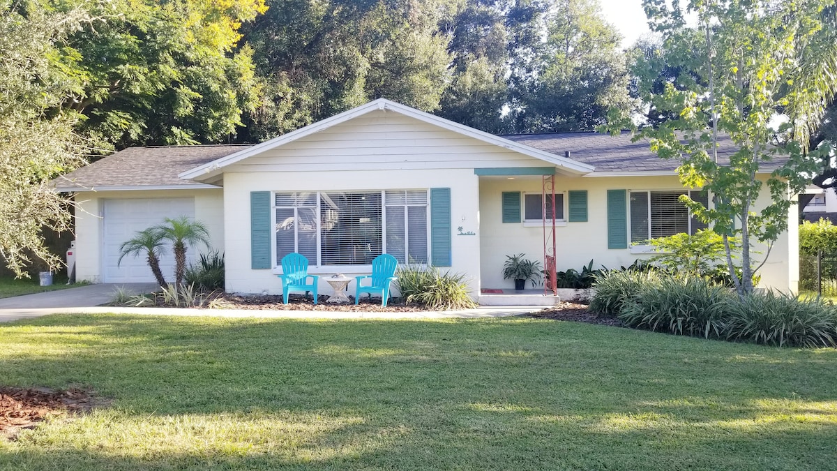 A Mid-Century cottage is presented with a well-kept exterior, featuring light yellow walls and turquoise shutters. A small seating area with two chairs is positioned on the front porch, surrounded by lush greenery, with a manicured lawn extending towards a tree-lined background.