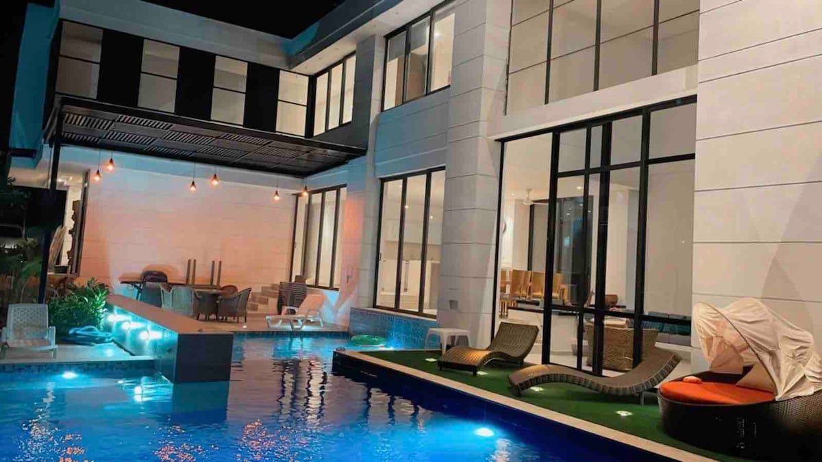 The exterior of the house is illuminated at night, showcasing a private pool with vibrant blue water and elegant lounge chairs nearby. Large glass windows provide a view into the spacious interiors, where comfortable seating areas are visible, creating an inviting atmosphere.