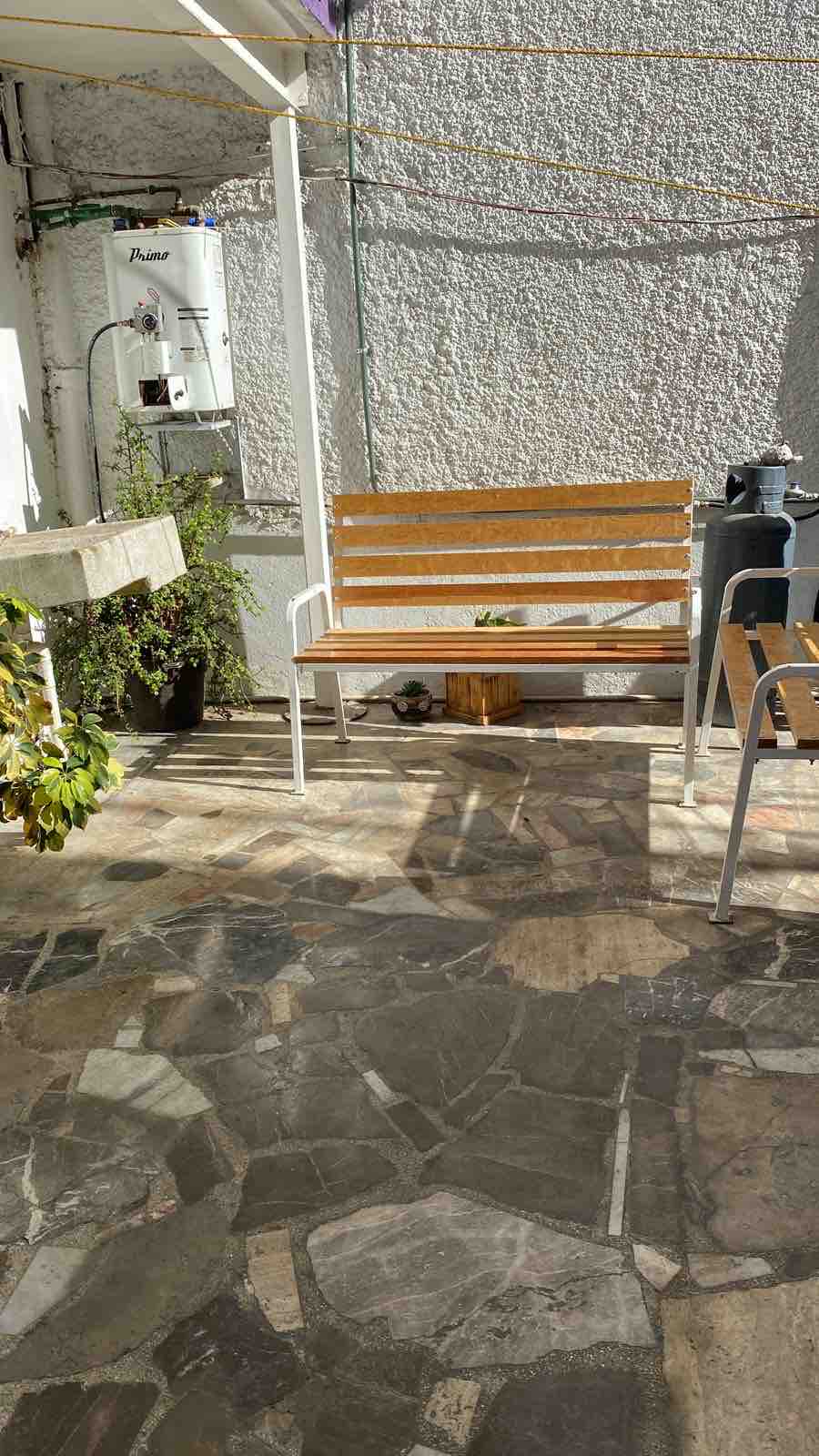 A small outdoor patio is depicted, featuring a wooden bench and a few metal chairs. A potted plant and a water heater are visible in the background. The floor is composed of uneven stone tiles, illuminated by natural sunlight.