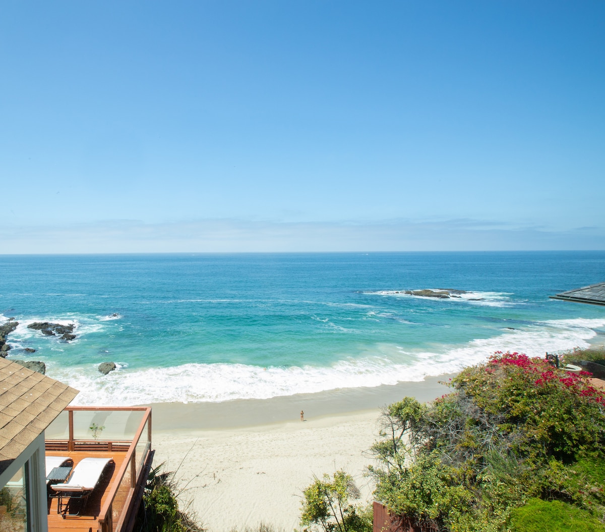 A serene ocean view captures a sandy beach bordered by gentle waves and a vibrant blue sky. The scene is complemented by lush greenery in the foreground, enhancing the natural beauty of the coastal setting.