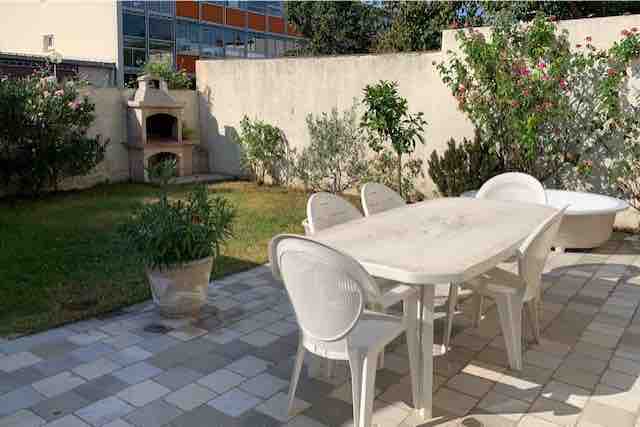 An outdoor garden space features a light-colored table surrounded by white chairs. A few potted plants add greenery, while a built-in stone barbecue is seen in the background. The area is bordered by low shrubs and a well-maintained lawn.