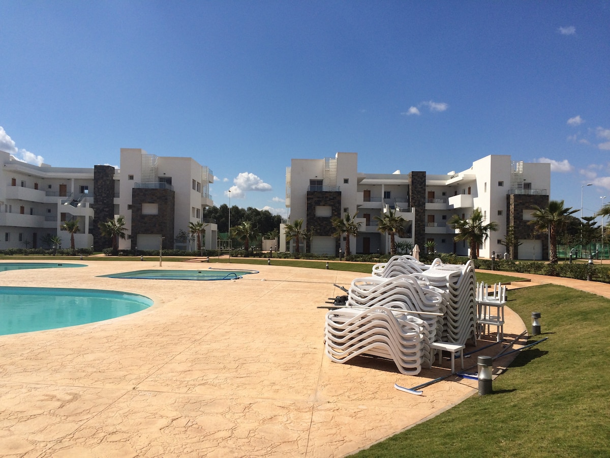 The image displays a sunny view of a residential complex featuring modern white buildings surrounded by a landscaped area. A large, inviting swimming pool is visible alongside stacked lounge chairs, with neatly trimmed grass and palm trees enhancing the outdoor space.