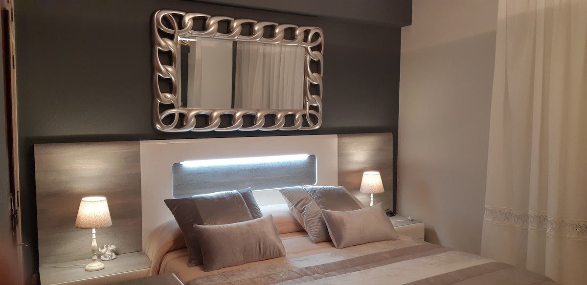 A well-appointed bedroom is presented with a spacious bed outfitted with neutral-toned linens. Two bedside lamps provide soft lighting, enhancing the tranquil ambiance. A large, decorative mirror hangs above the headboard, reflecting light from the nearby window, which is dressed with sheer white curtains.