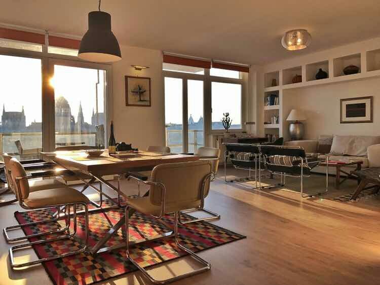 The living and dining area features large windows that offer river views, allowing for natural light to fill the space. A modern dining table is surrounded by stylish chairs, while a cozy seating area is positioned near the window, showcasing tasteful decor.