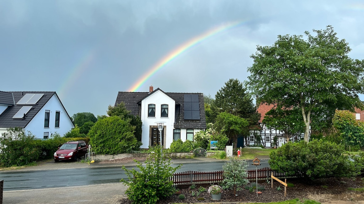 A suburban scene is depicted with a two-story house featuring a black roof and solar panels. Lush greenery surrounds the property, while a vibrant rainbow arcs across the sky. A red car is parked on the street, and a quaint garden is visible in the foreground.