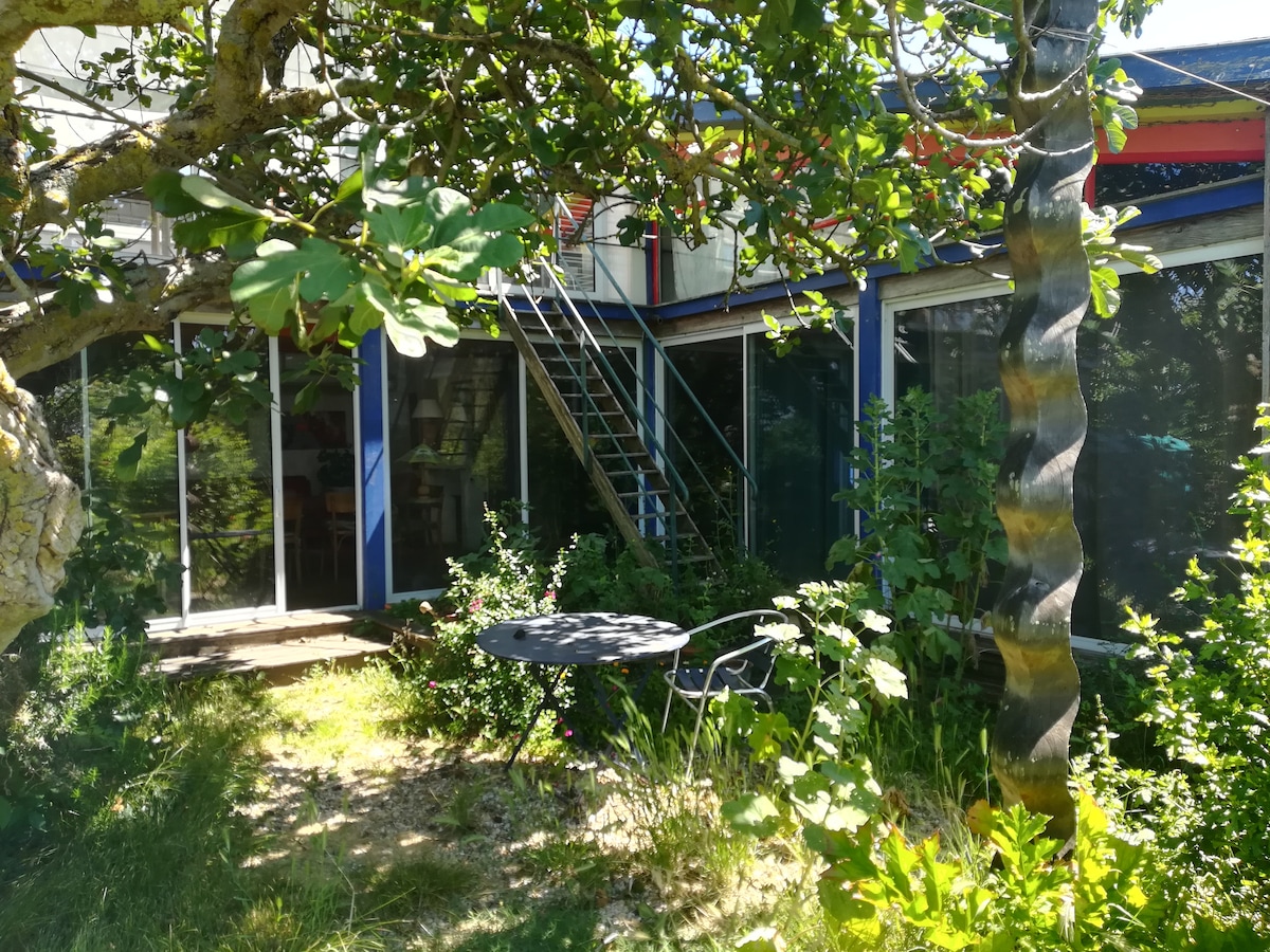 The exterior of a unique wooden house is surrounded by lush greenery. A small table with two chairs is positioned on a patio, shaded by trees. A set of stairs leads to a second level, with large glass doors reflecting the natural surroundings.