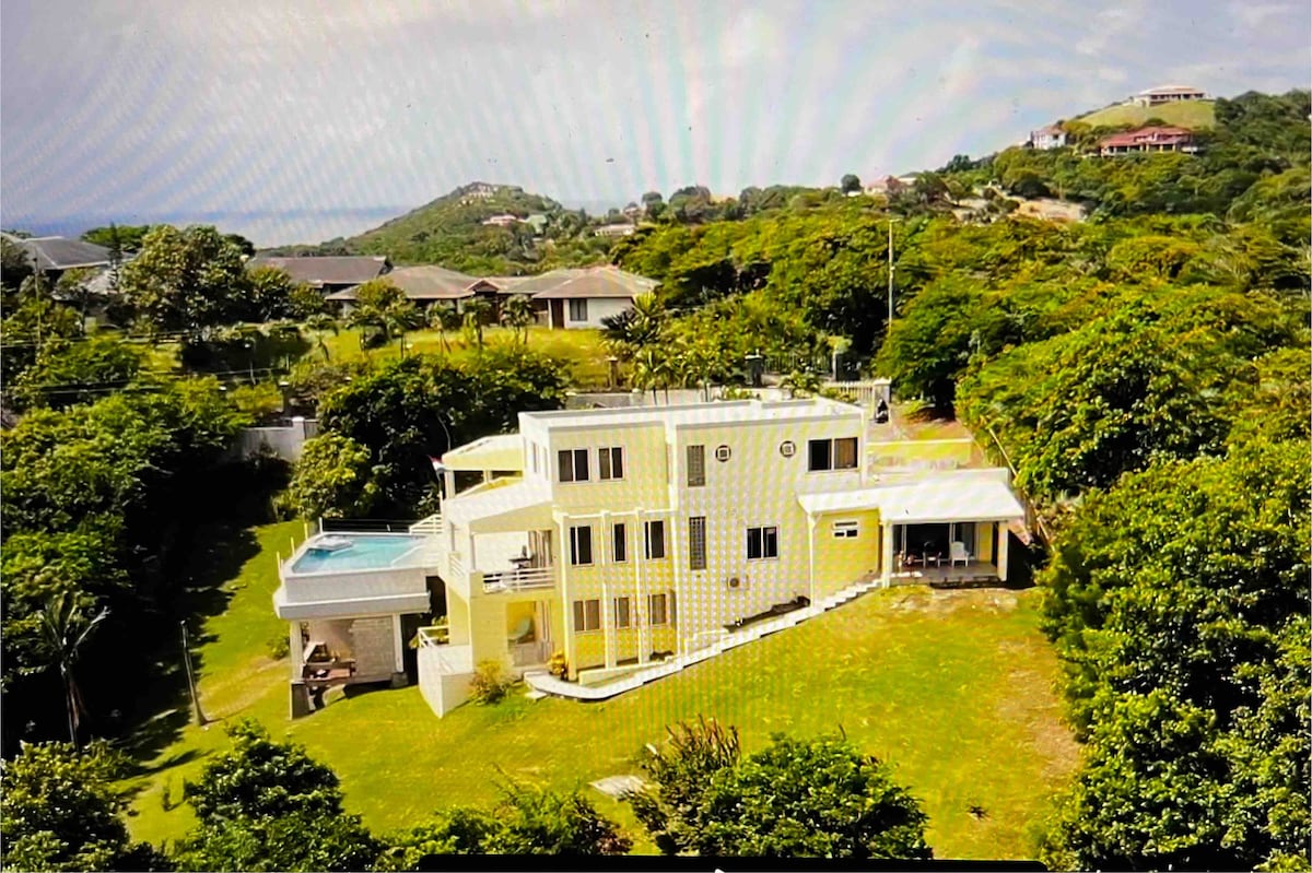 The exterior of the villa is presented against a backdrop of lush greenery and distant hills. A spacious lawn complements the yellow structure, which features multiple levels and large windows. A pool is visible on the upper terrace, surrounded by outdoor seating.