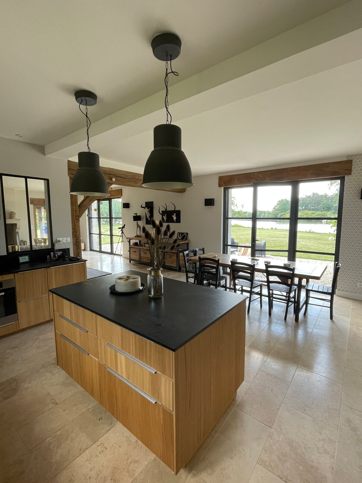 A modern kitchen features a central island with a dark countertop and natural wood cabinetry. Two large pendant lights hang overhead. Adjacent to the kitchen, a dining area is set with a table and several chairs, offering views of the outdoor landscape through expansive windows.