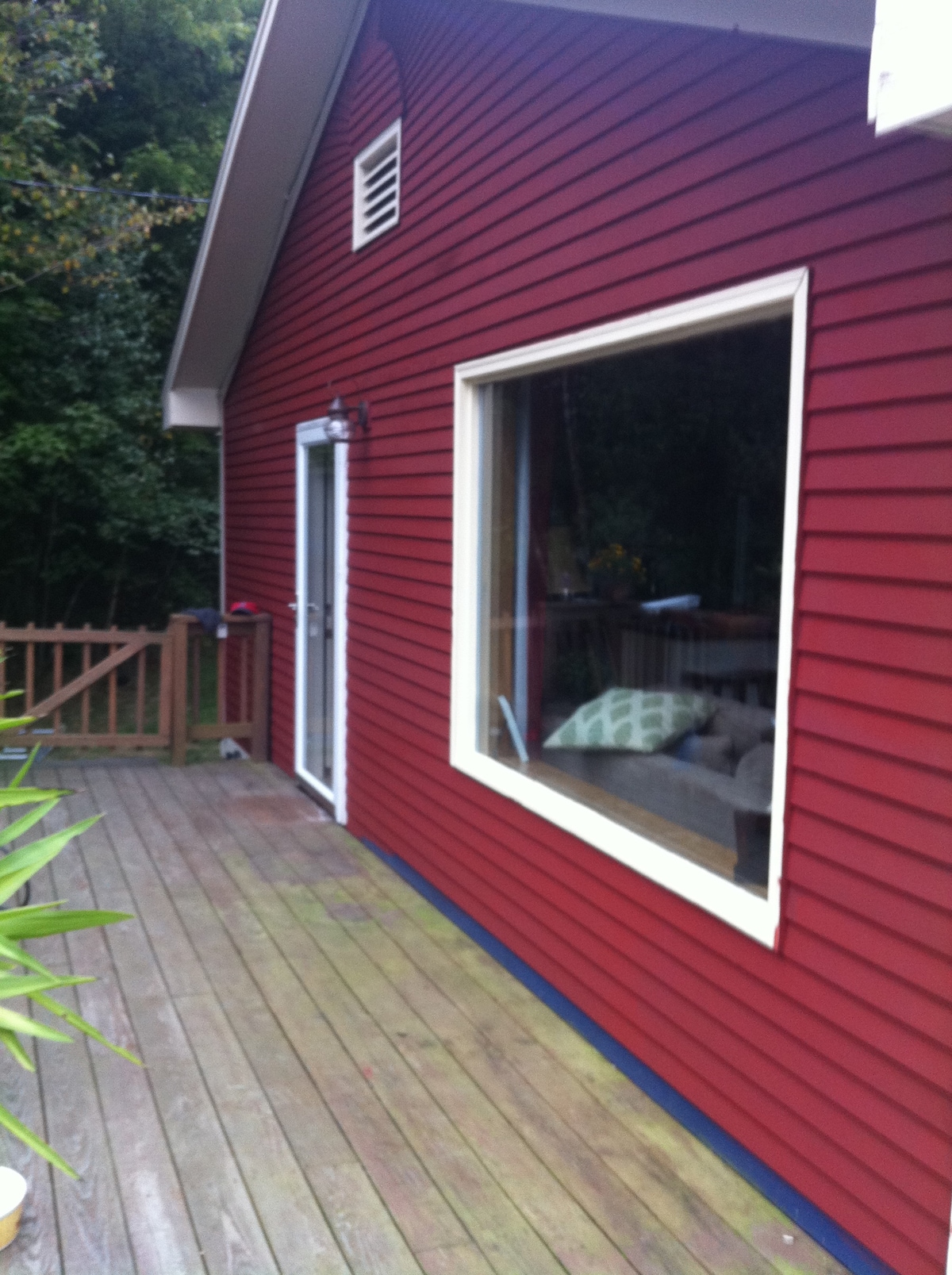 A cabin exterior is depicted, showcasing a red wooden siding and a large window reflecting natural surroundings. A wooden deck extends along the side, with a decorative railing and greenery visible in the background.