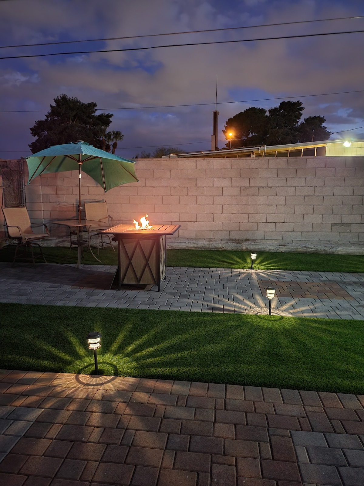 An outdoor patio area features a fire pit, surrounded by a circular seating arrangement. Soft lights cast shadows on the ground, and a large umbrella stands beside the fire pit. The grass is neatly trimmed, creating a welcoming space for relaxation.