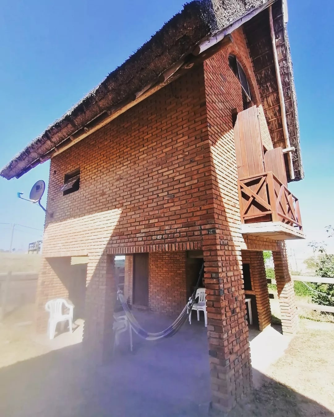 A charming two-story structure with a distinctive thatched roof is showcased. The brick exterior features a balcony with wooden railings and two white chairs positioned below. A hammock is suspended beneath the balcony, creating a relaxing outdoor space. The surrounding area appears open and inviting.