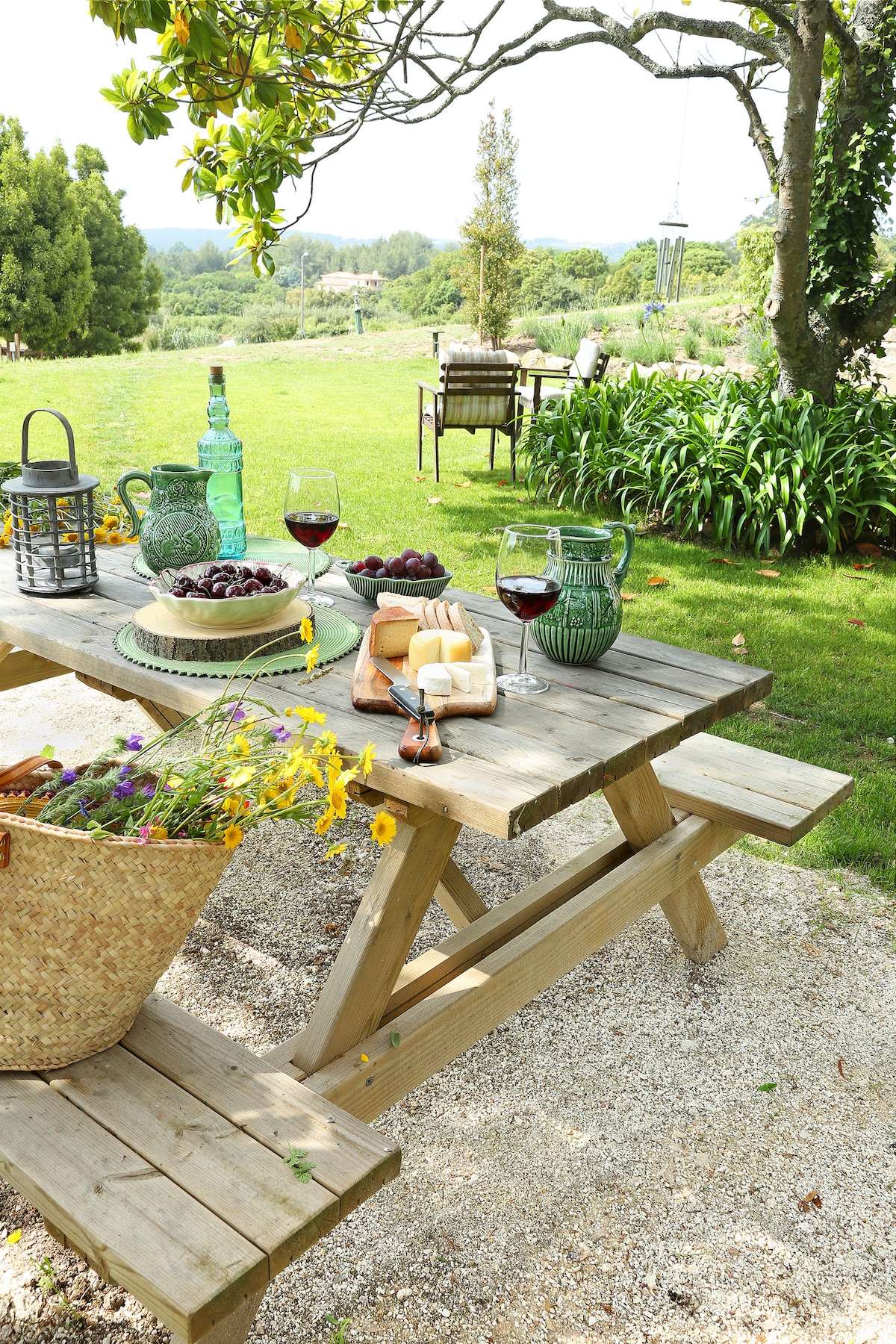 A wooden picnic table is set with cheese, fruit, and glasses of red wine. A basket of fresh flowers rests on the table. In the background, lush greenery and distant trees provide a scenic outdoor setting.