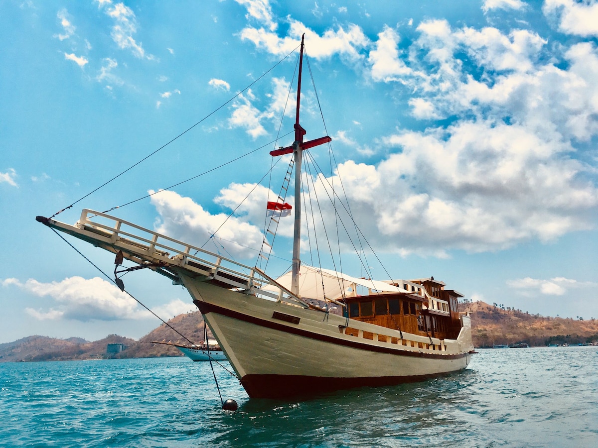 A traditional wooden boat is anchored in clear blue waters, with its sails partially extended. The boat features a spacious deck and multiple windows, surrounded by a scenic backdrop of rolling hills and a partly cloudy sky.